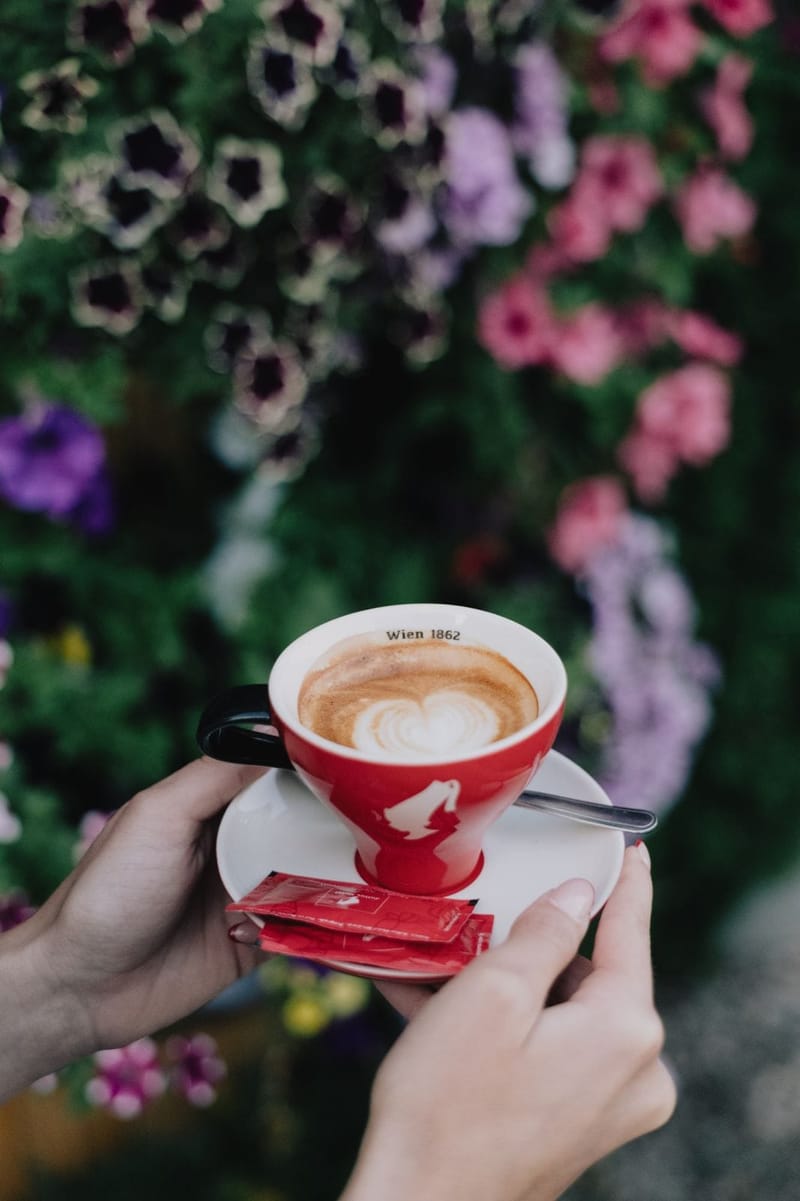 Cappuccino sa srcem od mlijeka u crvenoj šoljici, u rukama gošće, ispred cvijeća.